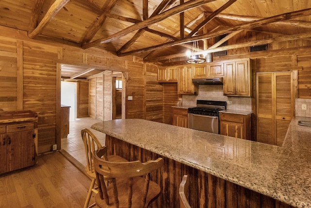 kitchen with vaulted ceiling with beams, wooden walls, under cabinet range hood, stainless steel gas range, and light wood finished floors