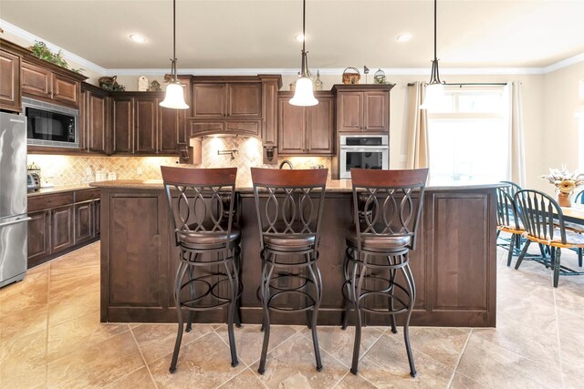 kitchen with dark brown cabinetry, hanging light fixtures, an island with sink, stainless steel appliances, and backsplash