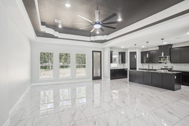 interior space featuring appliances with stainless steel finishes, decorative backsplash, a raised ceiling, ceiling fan, and a kitchen island with sink