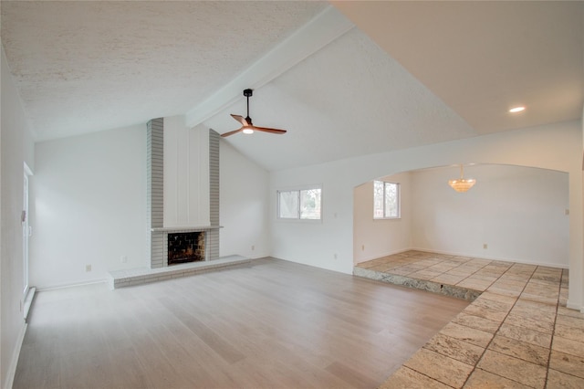 unfurnished living room featuring a brick fireplace, a textured ceiling, light hardwood / wood-style flooring, lofted ceiling with beams, and ceiling fan with notable chandelier