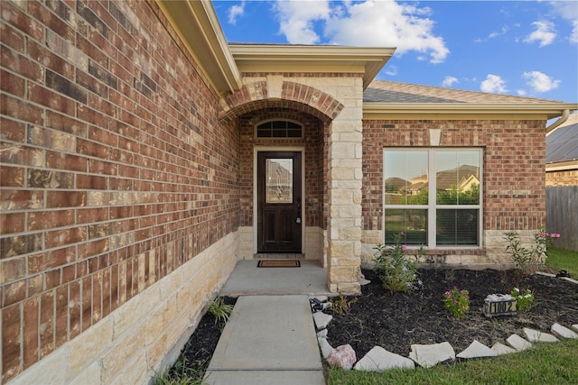 doorway to property featuring brick siding, roof with shingles, and fence