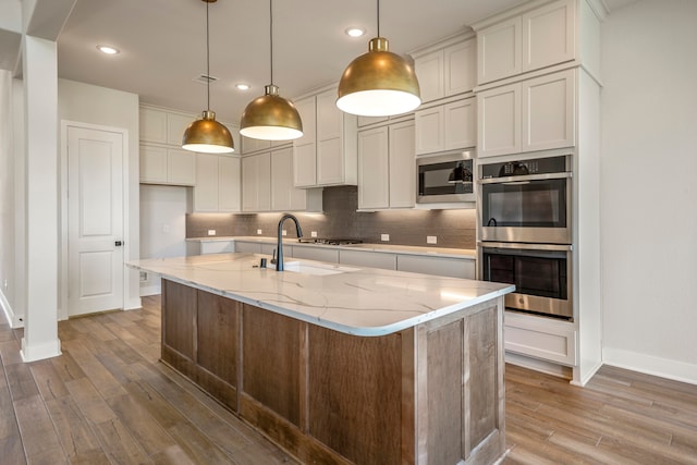 kitchen with stainless steel appliances, a center island with sink, decorative light fixtures, decorative backsplash, and hardwood / wood-style flooring