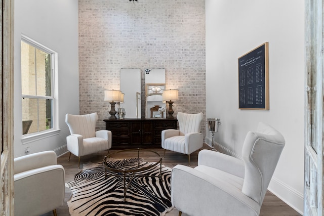 living area featuring a towering ceiling, wood-type flooring, a healthy amount of sunlight, and brick wall