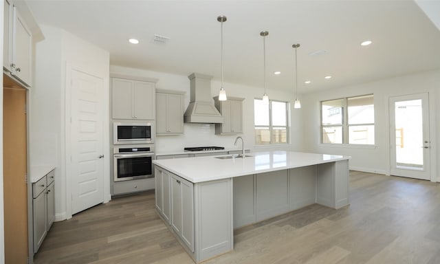 kitchen with premium range hood, stainless steel appliances, sink, a center island with sink, and hanging light fixtures