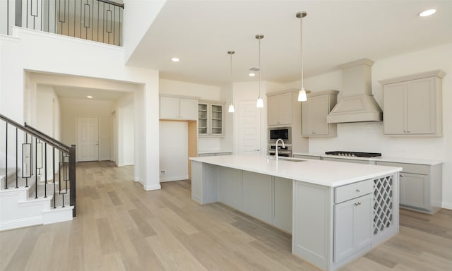 kitchen with gray cabinetry, custom exhaust hood, stainless steel appliances, a kitchen island with sink, and decorative light fixtures