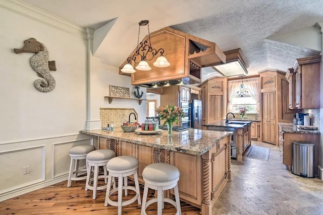 kitchen with a decorative wall, a peninsula, stainless steel fridge with ice dispenser, hanging light fixtures, and brown cabinets