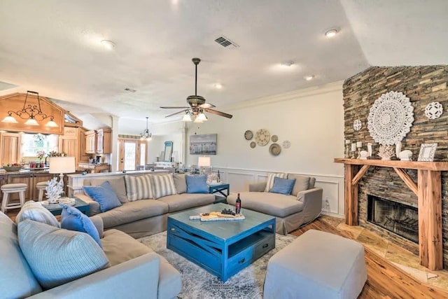 living area featuring lofted ceiling, a stone fireplace, visible vents, light wood-type flooring, and wainscoting