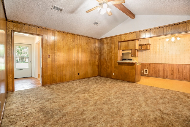 unfurnished living room with a textured ceiling, ceiling fan, light carpet, wood walls, and vaulted ceiling with beams