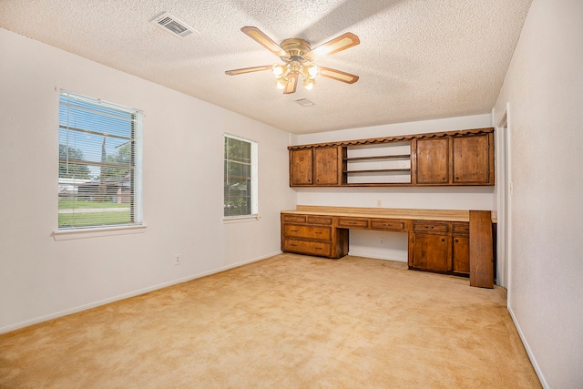 unfurnished office featuring ceiling fan, a textured ceiling, built in desk, and light carpet