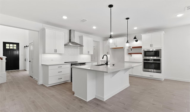 kitchen featuring sink, white cabinetry, an island with sink, oven, and wall chimney range hood