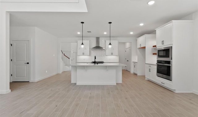 kitchen with built in microwave, an island with sink, sink, stainless steel oven, and wall chimney range hood