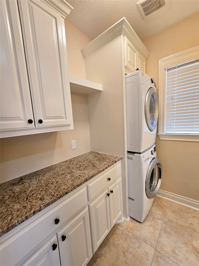 clothes washing area featuring a textured ceiling, light tile patterned floors, cabinets, and stacked washer and clothes dryer