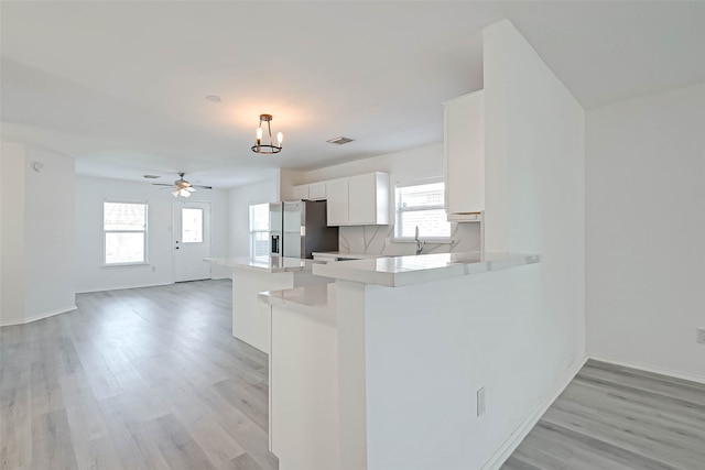 kitchen with kitchen peninsula, stainless steel refrigerator with ice dispenser, ceiling fan with notable chandelier, white cabinets, and light hardwood / wood-style floors