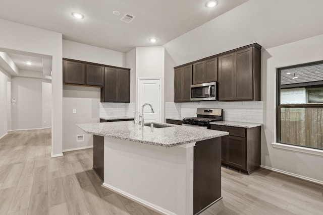 kitchen with stainless steel appliances, sink, light stone counters, light hardwood / wood-style floors, and an island with sink