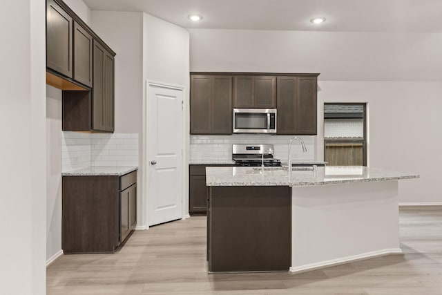 kitchen with light stone countertops, dark brown cabinets, an island with sink, and appliances with stainless steel finishes