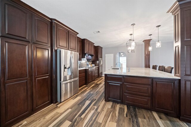 kitchen with dark brown cabinetry, sink, stainless steel refrigerator with ice dispenser, an island with sink, and dark wood-type flooring