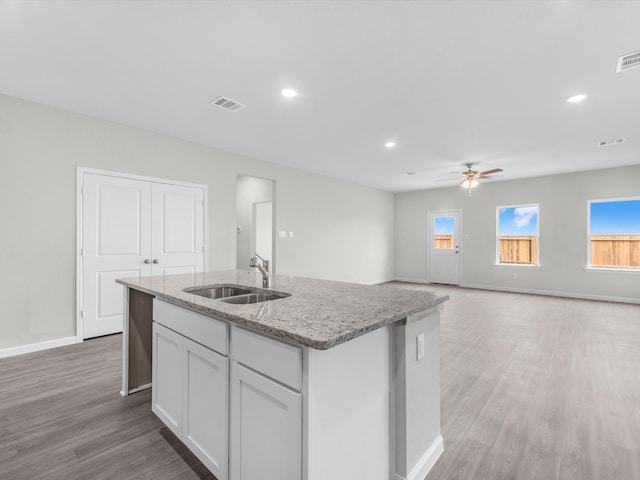 kitchen with light wood-type flooring, sink, light stone countertops, an island with sink, and ceiling fan