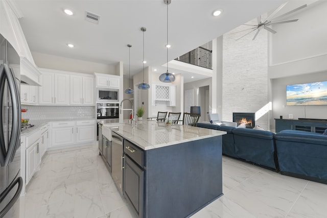 kitchen with appliances with stainless steel finishes, white cabinetry, light stone counters, a large island with sink, and a high ceiling