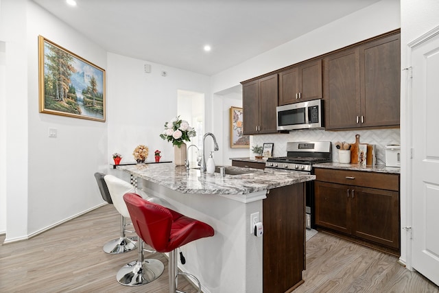 kitchen with an island with sink, appliances with stainless steel finishes, a breakfast bar, light wood-type flooring, and light stone counters