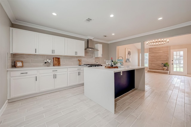 kitchen with crown molding, tasteful backsplash, wall chimney range hood, white cabinets, and an island with sink