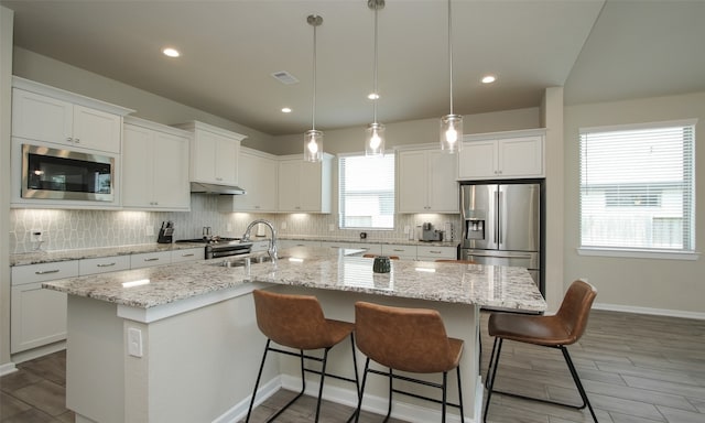 kitchen featuring stainless steel appliances, decorative backsplash, white cabinets, a center island with sink, and decorative light fixtures