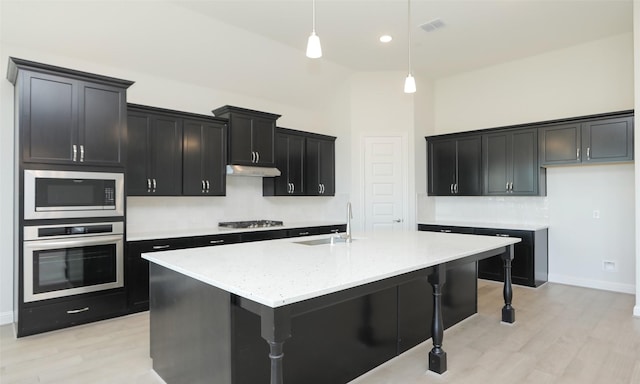 kitchen featuring a center island with sink, sink, light stone countertops, appliances with stainless steel finishes, and decorative light fixtures