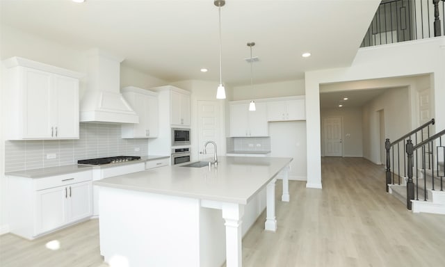 kitchen featuring custom range hood, stainless steel appliances, decorative light fixtures, a center island with sink, and white cabinetry