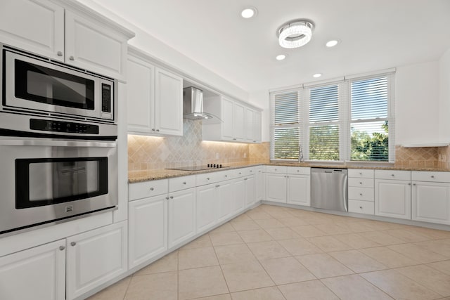 kitchen featuring decorative backsplash, wall chimney range hood, white cabinets, light tile patterned floors, and appliances with stainless steel finishes