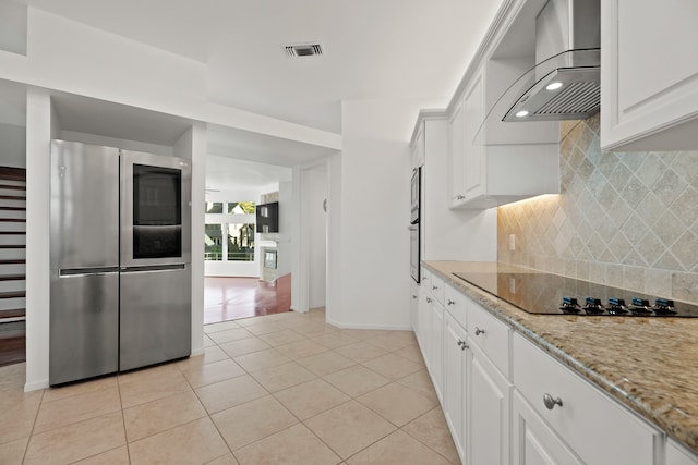 kitchen featuring wall chimney range hood, light stone counters, light tile patterned floors, white cabinetry, and stainless steel appliances