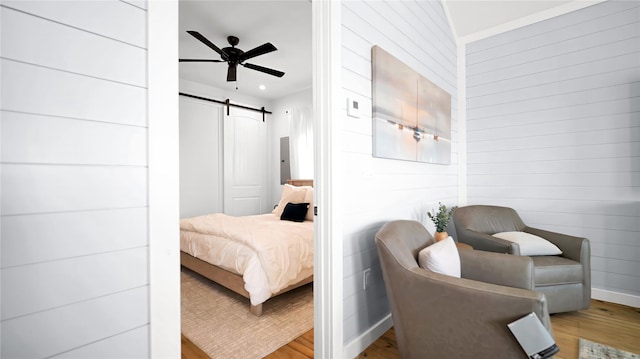 bedroom with a barn door, a ceiling fan, wooden walls, light wood-type flooring, and baseboards