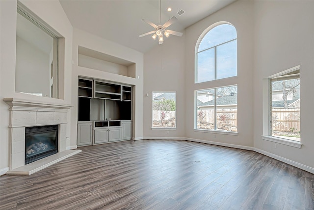 unfurnished living room with a high ceiling, hardwood / wood-style flooring, built in features, ceiling fan, and a tiled fireplace