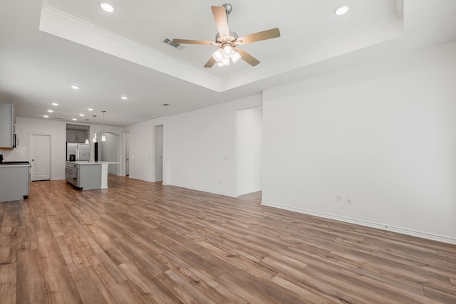 unfurnished living room with a raised ceiling, ceiling fan, ornamental molding, and light wood-type flooring