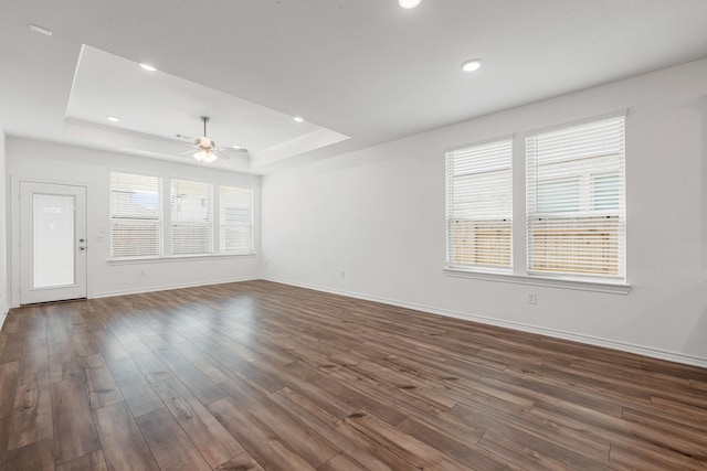 unfurnished living room featuring dark wood-type flooring, ceiling fan, a raised ceiling, and a healthy amount of sunlight