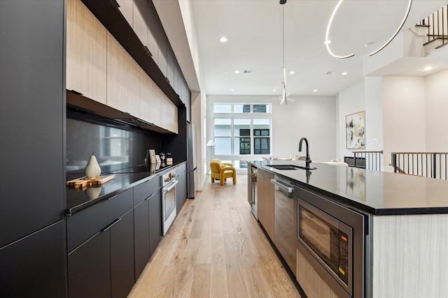 kitchen featuring appliances with stainless steel finishes, pendant lighting, an island with sink, sink, and light hardwood / wood-style flooring