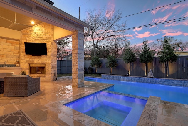 pool at dusk featuring pool water feature, an in ground hot tub, an outdoor stone fireplace, ceiling fan, and a patio