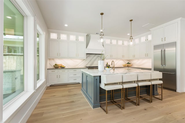 kitchen with white cabinetry, a kitchen breakfast bar, stainless steel built in refrigerator, an island with sink, and custom range hood