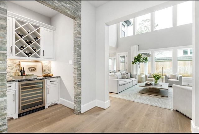 bar with dark stone counters, wine cooler, a towering ceiling, light hardwood / wood-style floors, and white cabinetry