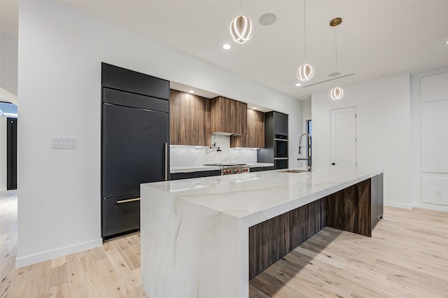 kitchen with paneled refrigerator, decorative light fixtures, light hardwood / wood-style floors, and an island with sink