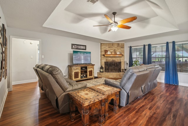 living room with a textured ceiling, dark hardwood / wood-style flooring, a brick fireplace, ceiling fan, and a tray ceiling