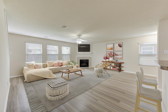 living room with light wood-type flooring, a tiled fireplace, ceiling fan, and a textured ceiling