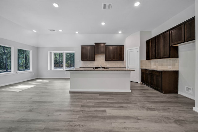 kitchen featuring an island with sink, backsplash, dark brown cabinetry, light stone counters, and light hardwood / wood-style floors