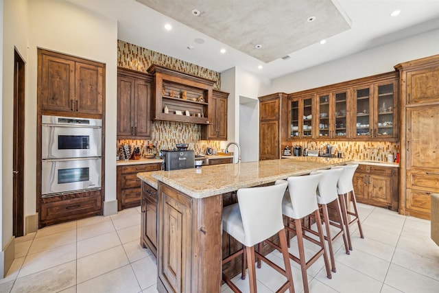 kitchen with light stone counters, light tile patterned floors, an island with sink, and stainless steel double oven