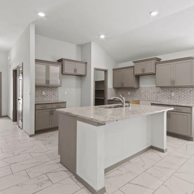 kitchen with sink, light stone counters, vaulted ceiling, gray cabinets, and a kitchen island with sink