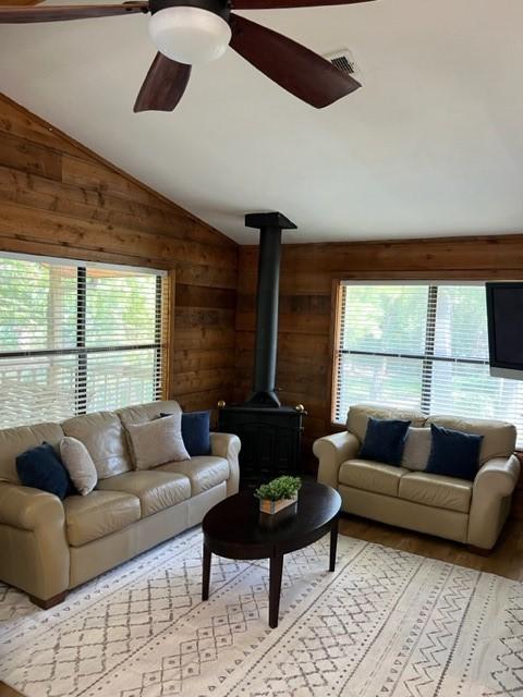 living room featuring wood walls, lofted ceiling, a wood stove, light hardwood / wood-style flooring, and ceiling fan