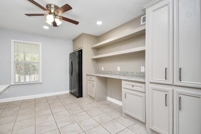 kitchen with light tile patterned floors, built in desk, light stone countertops, ceiling fan, and stainless steel fridge