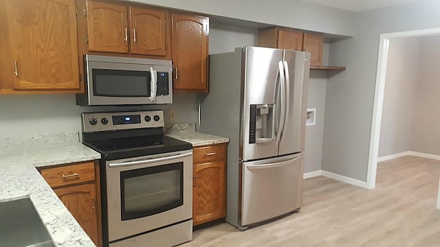 kitchen with brown cabinetry, stainless steel appliances, light wood-style flooring, baseboards, and light stone countertops