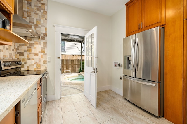 kitchen featuring wall chimney exhaust hood, brown cabinetry, tasteful backsplash, appliances with stainless steel finishes, and baseboards