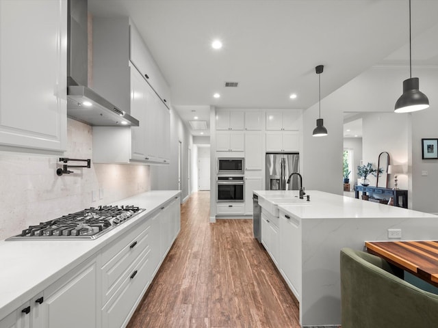 kitchen with stainless steel appliances, white cabinets, a sink, wall chimney range hood, and wood finished floors