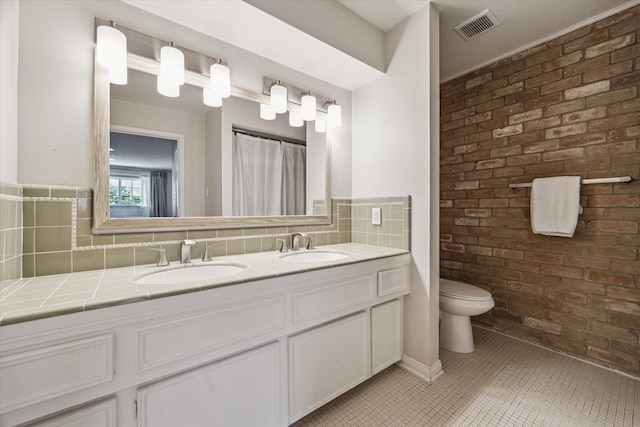 bathroom with tile patterned floors, tasteful backsplash, double vanity, brick wall, and toilet