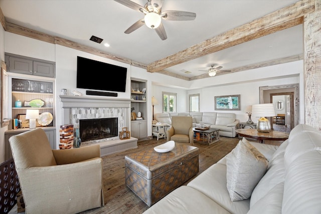 living room with built in features, a fireplace with raised hearth, beamed ceiling, a ceiling fan, and wood finished floors